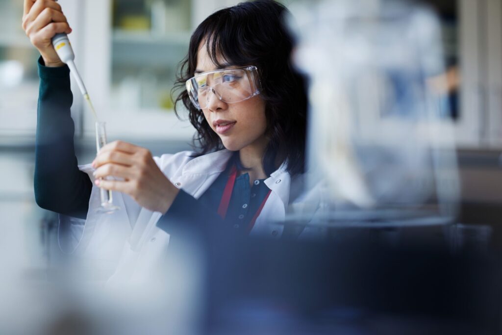 A woman wearing safety goggles and a lab coat uses a pipette to transfer liquid to a test tube in a lab.